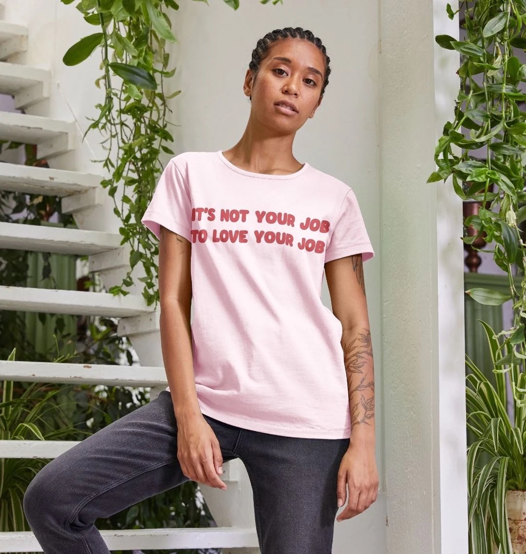 Model wearing a light pink t-shirt with a large dark pink text along the front on two lines reading It's Not Your Job To Love Your Job.